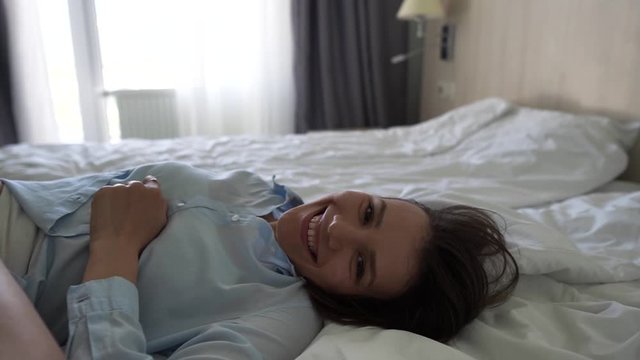 A young happy girl in a blue shirt lies on a bed in a hotel room and smiles