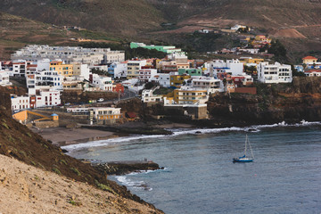 Sardina de Galdar small town on the coast with sailboat on calm sea in Gran Canaria, Spain. Colorful houses of village in Canary Islands at sunset. Dark tones effect applied