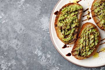 Flat lay of freshly made minced avocado toast with balsamico dressing, linseed or flaxseed and oil on a white plate, on a gray background