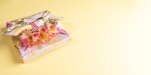 Gift box trendy wrapped in floral cloth in japanese Furoshiki technique with flowers Alstroemeria on yellow background. Valentine's day or Women's day festive banner. Zero Waste. Top view, copy space.