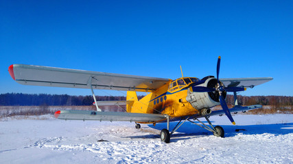 A yellow old biplane plane is parked on a winter airfield against a background of bright blue sky...