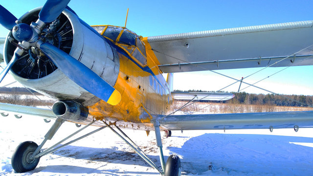 A yellow old biplane plane is parked on a winter airfield against a background of bright blue sky and white snow with the engine running and smoke