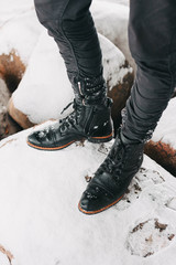 male legs in black cool leather lace-up boots stand in the snow on large stones in winter