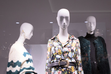 three or a group of female bald white mannequins in a shop window. Mannequins are dressed in dresses, sweaters, a fur coat, jewelry.