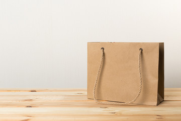 An empty paper bag with handles for shopping, on the front right side of a table in light wood with a light wall background, providing copy space on the left.