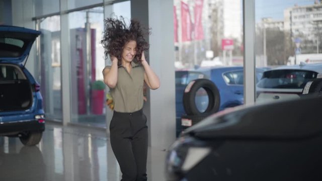 Slow motion of excited Caucasian young woman turning to new car and jumping up. Happy woman with curly hair buying vehicle in car dealership. Automobile industry, success, purchasing.