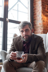 Portrait of brutal middle-aged man in jacket sitting in comfortable armchair and drinking whisky alone