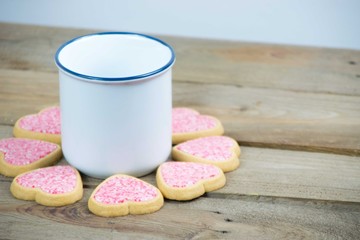 White and blue ceramic mug surrounded by heart shaped cookies with pink frosting and sprinkles on a wooden board. Love concept