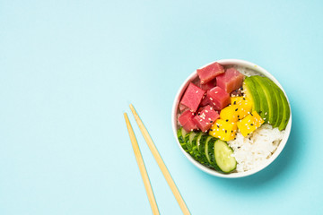 Tuna poke bowl with rice, avocado, mango and cucumber on blue.