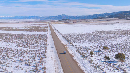 AERIAL: Scenic asphalt highway leads silver SUV across the snowy landscape.