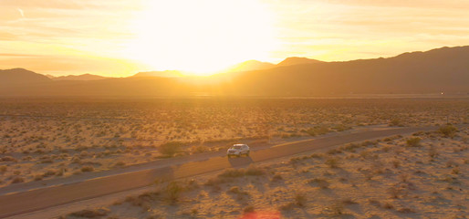 AERIAL: Cinematic shot of golden sun rays shining on SUV crossing Nevada desert.