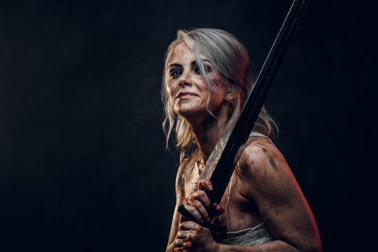 Naked Fantasy woman warrior wearing rag cloth stained with blood and mud holding a sword and smiling. Studio photo on a dark background. Cosplayer as Ciri from The Witcher