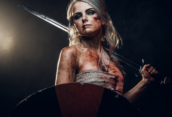 Naklejka premium Fantasy woman warrior wearing rag cloth stained with blood and mud, holding sword and shield. Studio photo on a dark background. Cosplayer as Ciri from The Witcher
