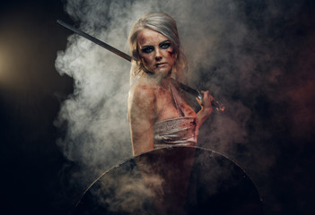 Woman warrior wearing rag cloth stained with blood and mud posing with a sword and shield. Studio...