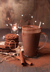 A transparent cup of hot chocolate with cinnamon on a wooden table with oatmeal cookies and chocolate. Against the background of lights.