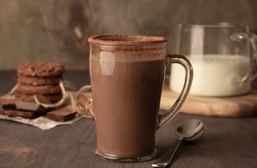 Transparent cup of hot chocolate with cinnamon and milk on a wooden table with oatmeal cookies and chocolate.