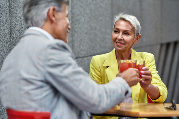 Time to relax. Happy beautiful middle-aged woman in yellow suit jacket drinking cocktails with her friend and smiling while sitting in cafe outdoors together