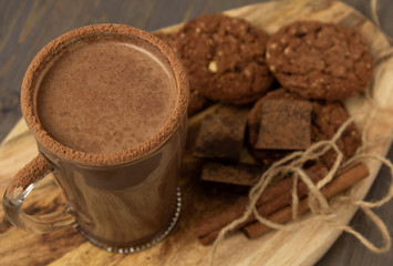 Transparent cup of hot chocolate with cinnamon and milk on a wooden board with oatmeal cookies and chocolate.