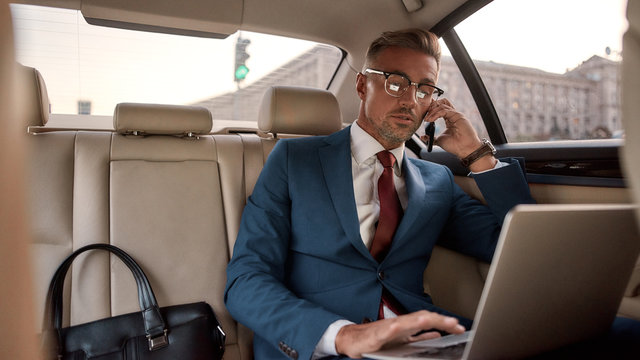 Working Anywhere. Confident and stylish mature businessman in full suit working on his laptop and talking on the phone with client while sitting in the car
