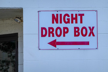 White cinder block wall with a white sign with red letters that say night drop box with an arrow