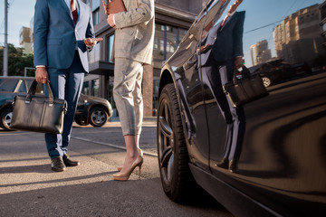 Cropped photo of two business people in classic wear standing near black car outdoors