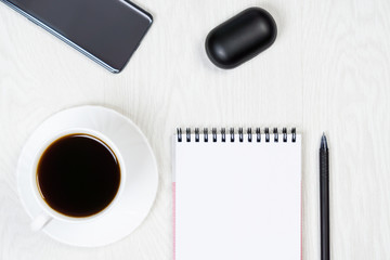 White cup of coffee, notebook, pen, phone, headphones on a white table