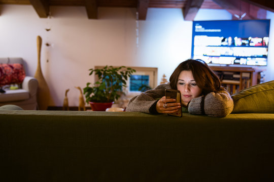 Cute young woman looking at phone in her hand shopping online while sitting on the couch in beautiful interior living room, television on background, relaxation and buying online icon 