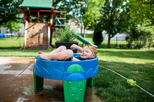 Toddler boy smirking as he lays in water table with clothes on outside