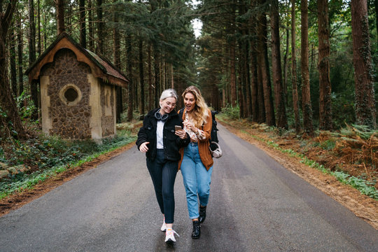 Two women walking down the road looking at their phone and laughing - Powered by Adobe