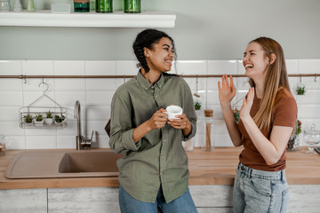 Girls teenagers talking on kitchen and drinking coffee, laughing and having funny joyful mood. International friendship, happy time together, friends having relax time. Enjoy communication and holiday