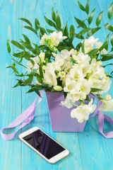 A delicate bouquet of white freesia in a lilac basket and a smartphone on a wooden table. Spring