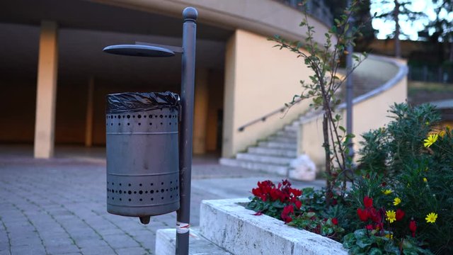 Black metal trash can in public park near the flowerbed, clean and empty garbage bin on the street. Person's awareness, saving world planet and preventing from global pollution