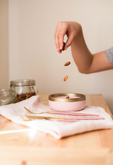 A healthy breakfast, a female hand pours nuts into a pink plate from a jar, and nearby are cutlery of gold on a napkin. Wooden table.
