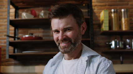 Obraz premium Portrait of sexy handsome mature man in shirt smiling on camera standing in his stylish kitchen relaxing at home apartment on the weekend.