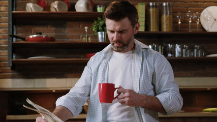 Handsome man in shirt sipping morning coffee reading news on a newspaper relaxing at breakfast in his luxury home kitchen.