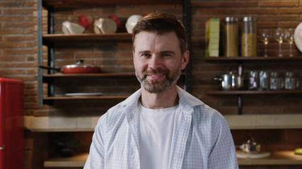Portrait of a mature handsome bearded man in white shirt standing by kitchen table and smiling of...