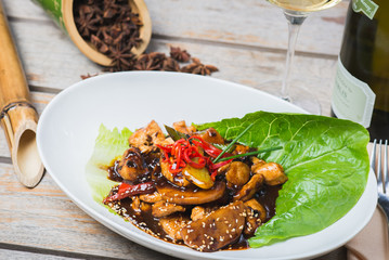 Sliced chicken fried in sweet and sour soy sauce with vegetables and herbs in a deep plate on a wooden table in a restaurant.