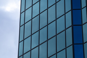 The exterior glass wall of a tall sky scraper building. The windows are blue in color in the foreground with blue sky and clouds in the background. The sky is reflecting in the building's windows. 