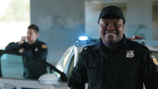 Happy african american young man cops stand near patrol car look at camera smile enforcement officer police uniform auto safety security communication control policeman portrait close up slow motion