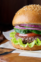 Burger on wood board at dark background. Sesame bun with beef, lettuce, tomato, cucumber and onion