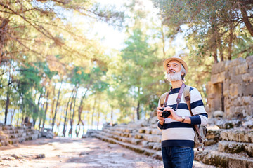 Enjoying travel. Senior smiling man with backpack holding camera on ancient sightseeing background.