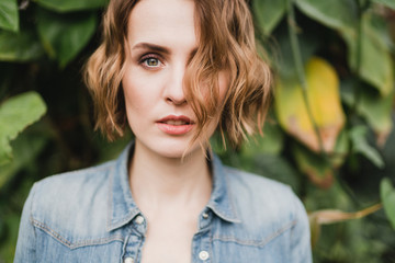 Fototapeta premium Young beautiful caucasian woman in glass greenhouse among colorful greenery leaves and flowers. Art portrait of a girl wearing a shirt. Gardener, nature lover, inspiration concept.