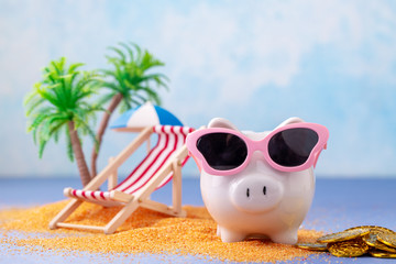 Piggy bank with sunglasses on miniature beach with deckchair, palms and umbrella on blue background...