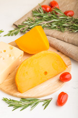 Various types of cheese with rosemary and tomatoes on wooden board on a white wooden background. Side view, close up.