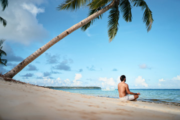 Fototapeta na wymiar Suntan and meditation. Enjoying vacation.. Relaxed young man siiting on lotus position on the tropical beach.