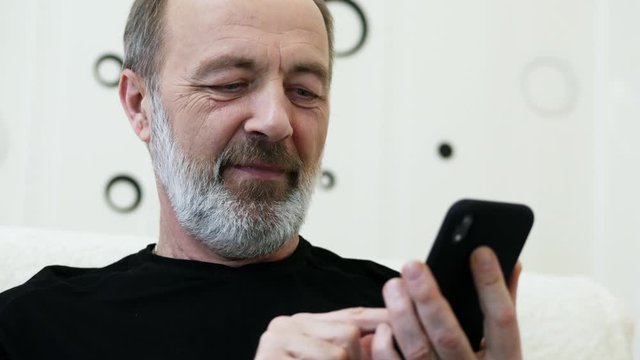 Middle-aged man with a gray beard uses the mobile phone at home, browsing internet in close-up