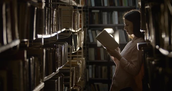 Young pretty female looking through book in dark library storage room, searching information