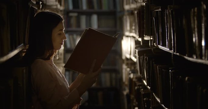 Young pretty female student reading book in dark library room, interesting information