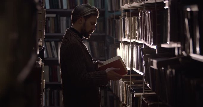 Handsome middle-aged bearded male looking through dusty book in library room, old edition