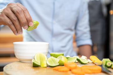 Asian Thai people holds and squeezes green lime (lemon) on the top of white little bowl with quater limes on wood plate. The environment is simple wood kitchen theme.